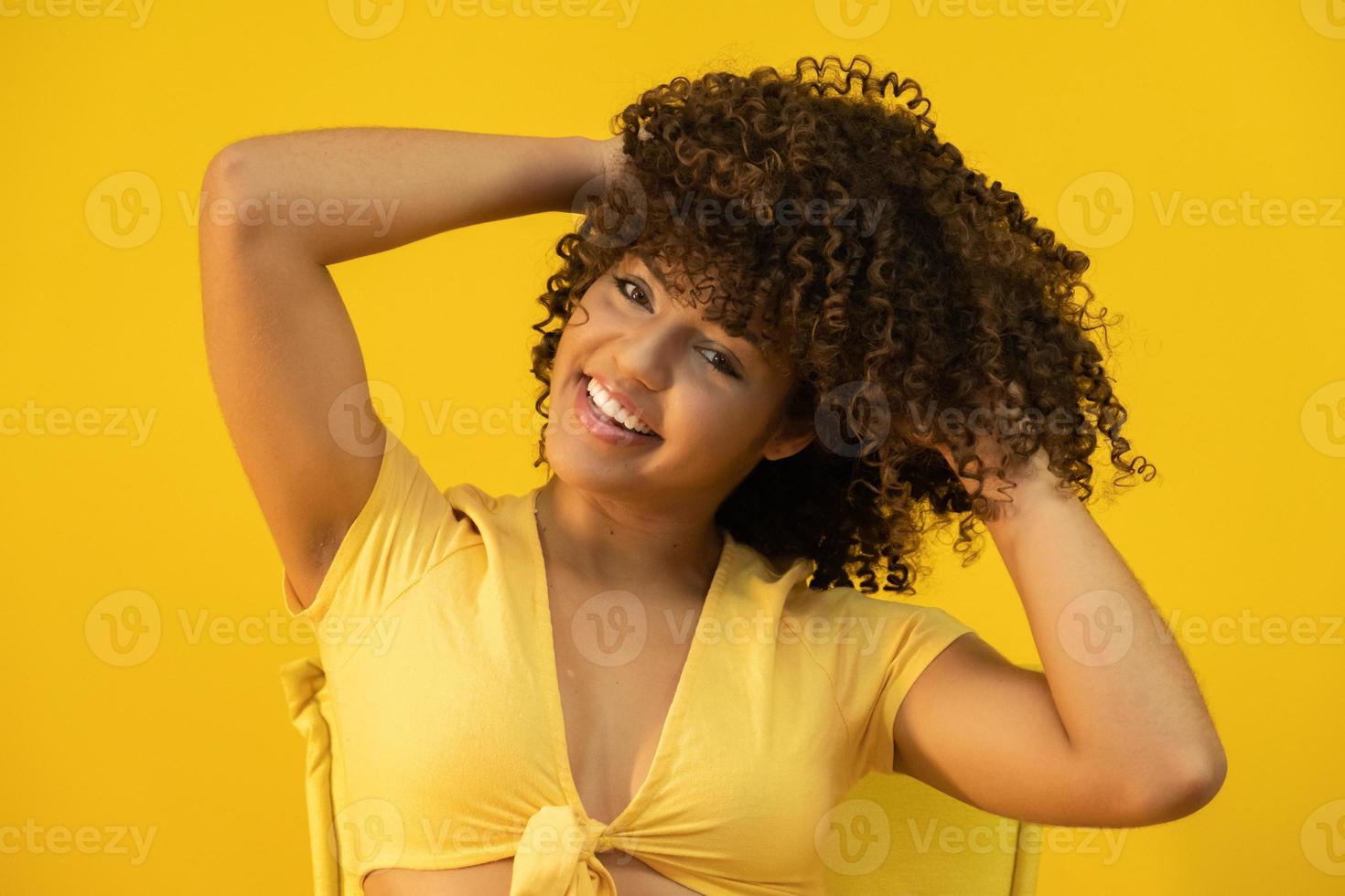 heureuse femme africaine américaine qui rit avec ses cheveux bouclés sur fond jaune. femme bouclée riante en pull touchant ses cheveux et regardant la caméra. photo