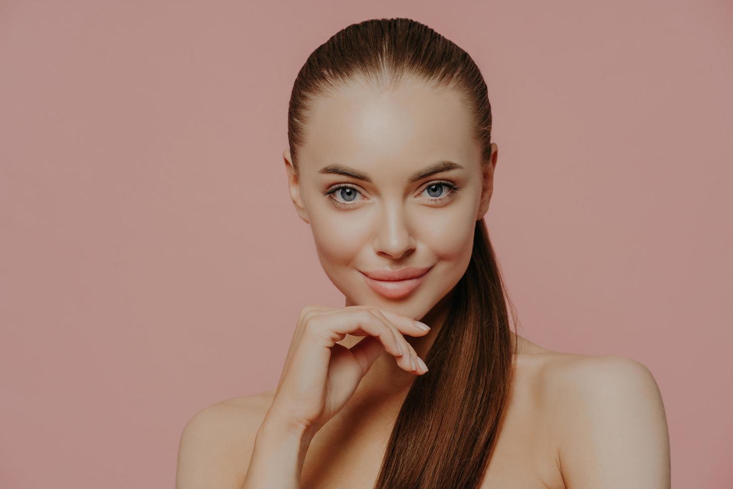 soins de la peau et procédures anti-âge. une jeune femme brune confiante garde la main sous le menton, a les cheveux peignés, une peau propre et fraîche, bénéficie de soins de beauté, pose avec un corps nu sur fond rose photo