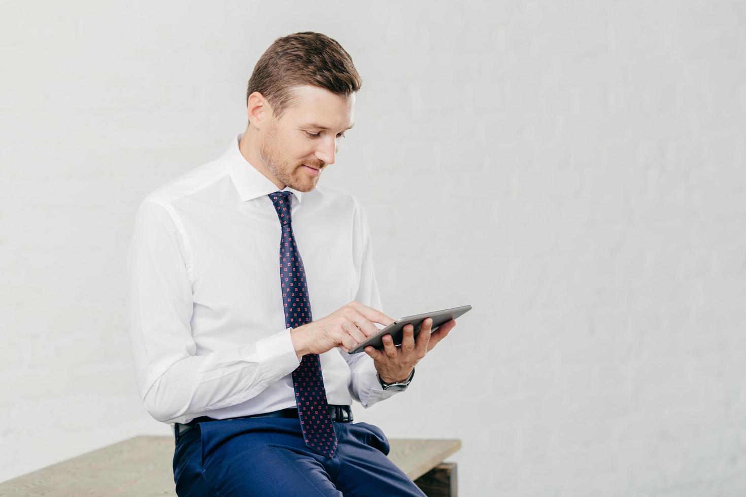 photo horizontale d'un jeune homme d'affaires prospère qui met à jour son profil sur un blog d'entreprise utilise une application sur un ordinateur tablette, porte des vêtements élégants, isolé sur fond blanc dans un immeuble de bureaux