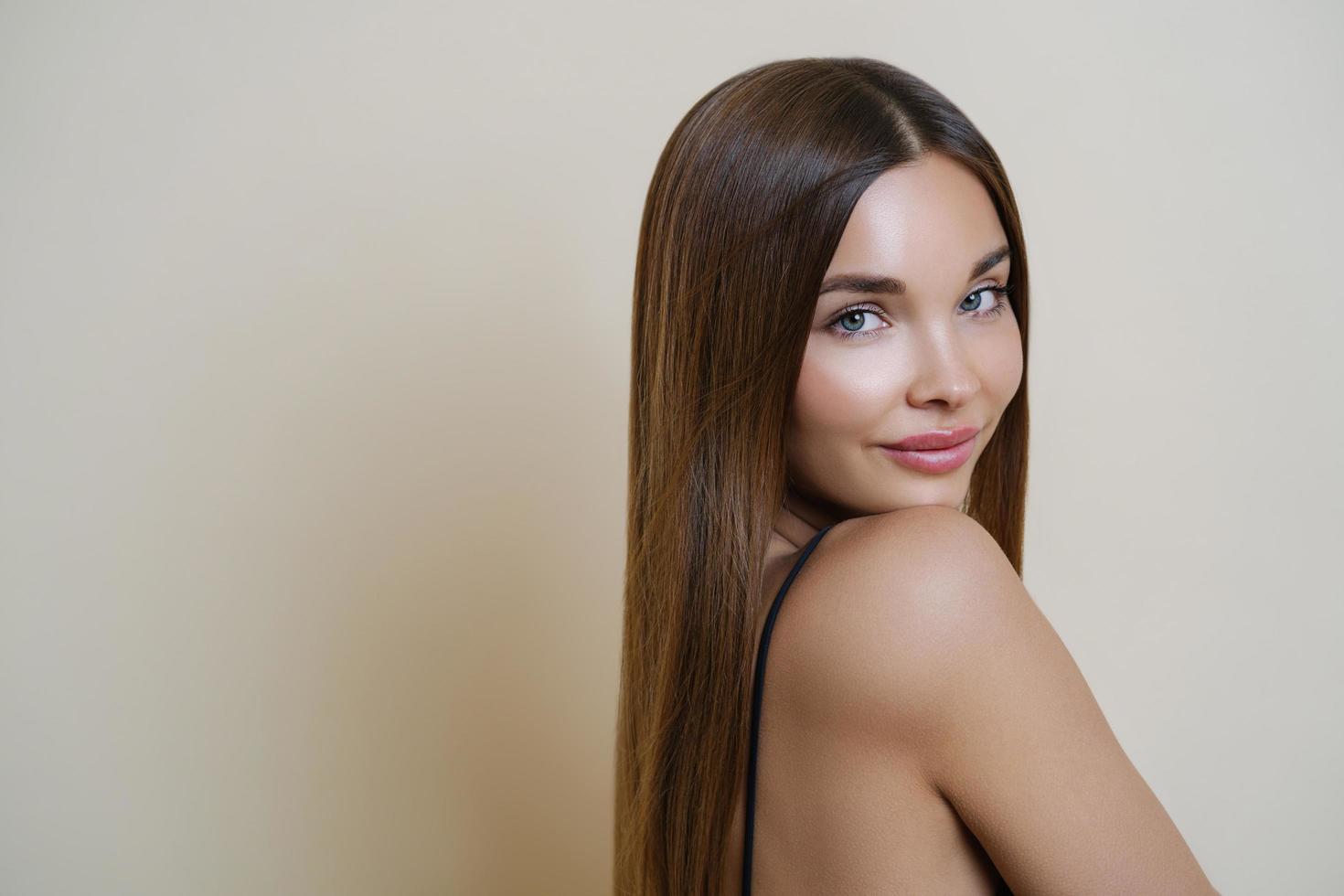 séduisante jeune femme européenne avec une peau douce et saine, un corps parfait, semble calme directement à la caméra, a les cheveux longs, se tient à l'intérieur sur fond beige. concept jeunesse, beauté et soins de la peau. photo