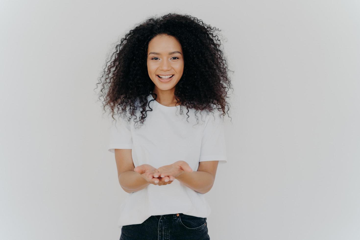 heureuse femme à la peau sombre a les mains en coupe, demande à donner quelque chose, sourit agréablement, a les cheveux crépus touffus, porte un t-shirt et un jean, isolé sur fond blanc. concept de personnes et de charité photo