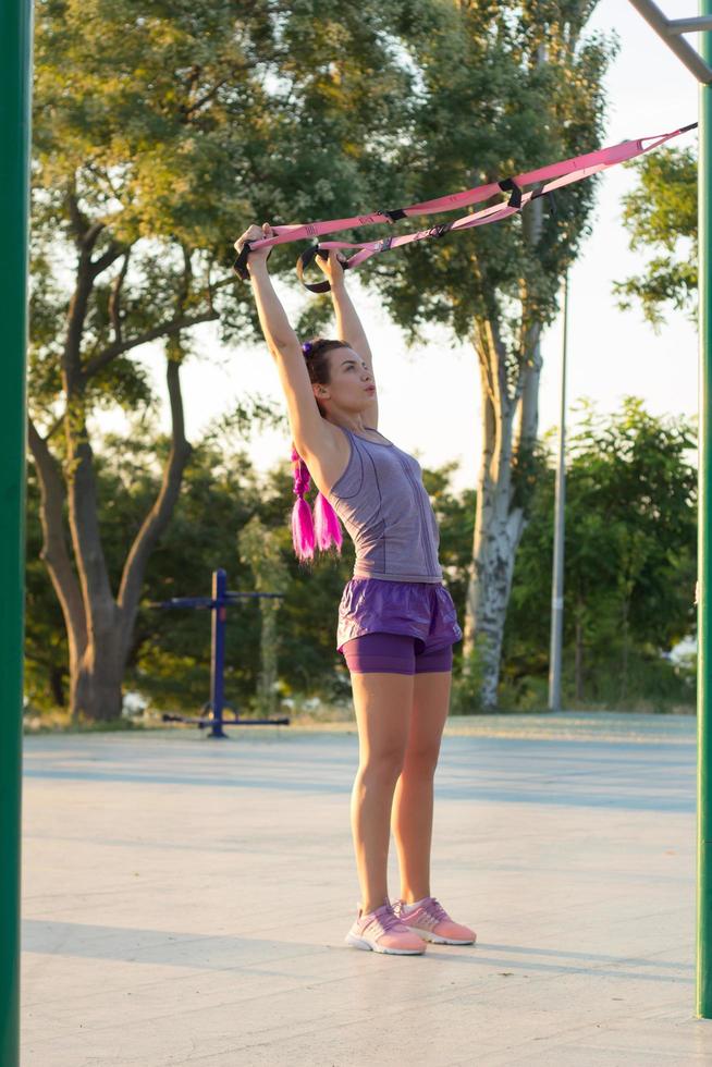 belle femme en forme de sportwear rose et violet s'entraînant sur une salle de sport en plein air le matin, exercices avec des sangles de suspension dans le parc photo