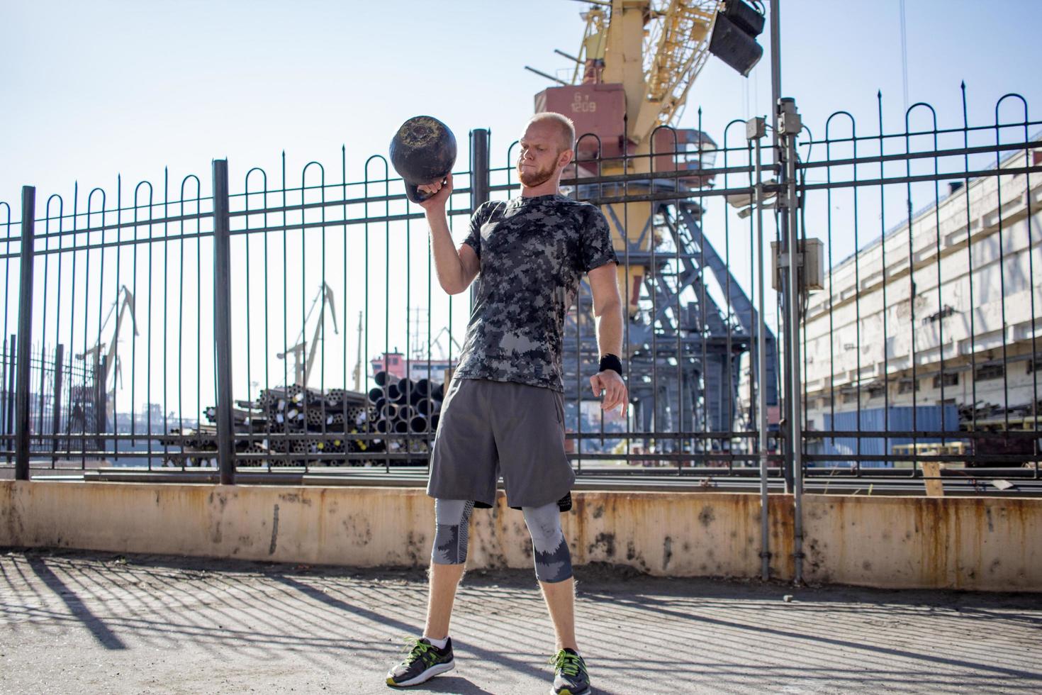 jeune athlète masculin barbu s'entraînant dans une zone industrielle par temps ensoleillé, exercices de kettlebells à l'extérieur, arrière-plan urbain photo