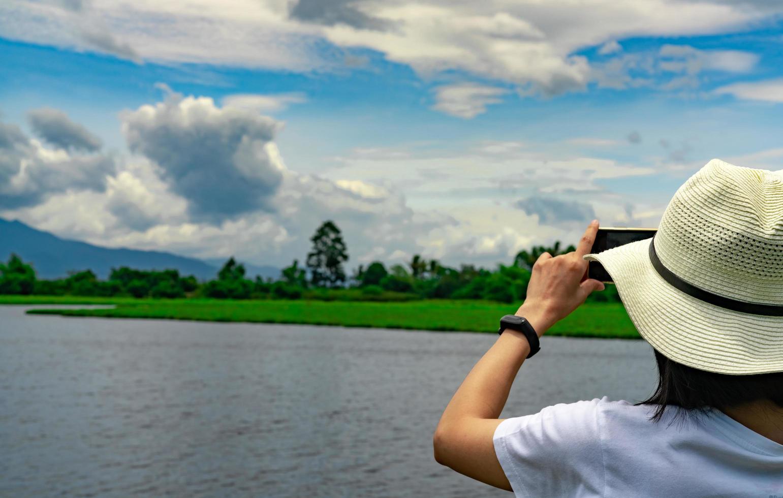 une femme asiatique utilise un smartphone pour prendre une photo d'un magnifique paysage de rivière, de montagne et de forêt. voyage de vacances d'été. femme hipster avec une activité de plein air de style minimal pour une vie relaxante. voyager seul.