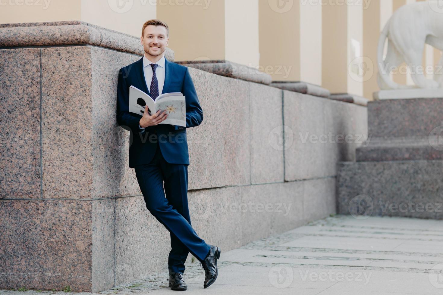 la photo d'un patron masculin qui a réussi se tient les jambes croisées, tient un magazine d'affaires, lit les informations nécessaires, se prépare à la présentation devant des collègues, se tient près d'un immeuble de bureaux, s'habille formellement