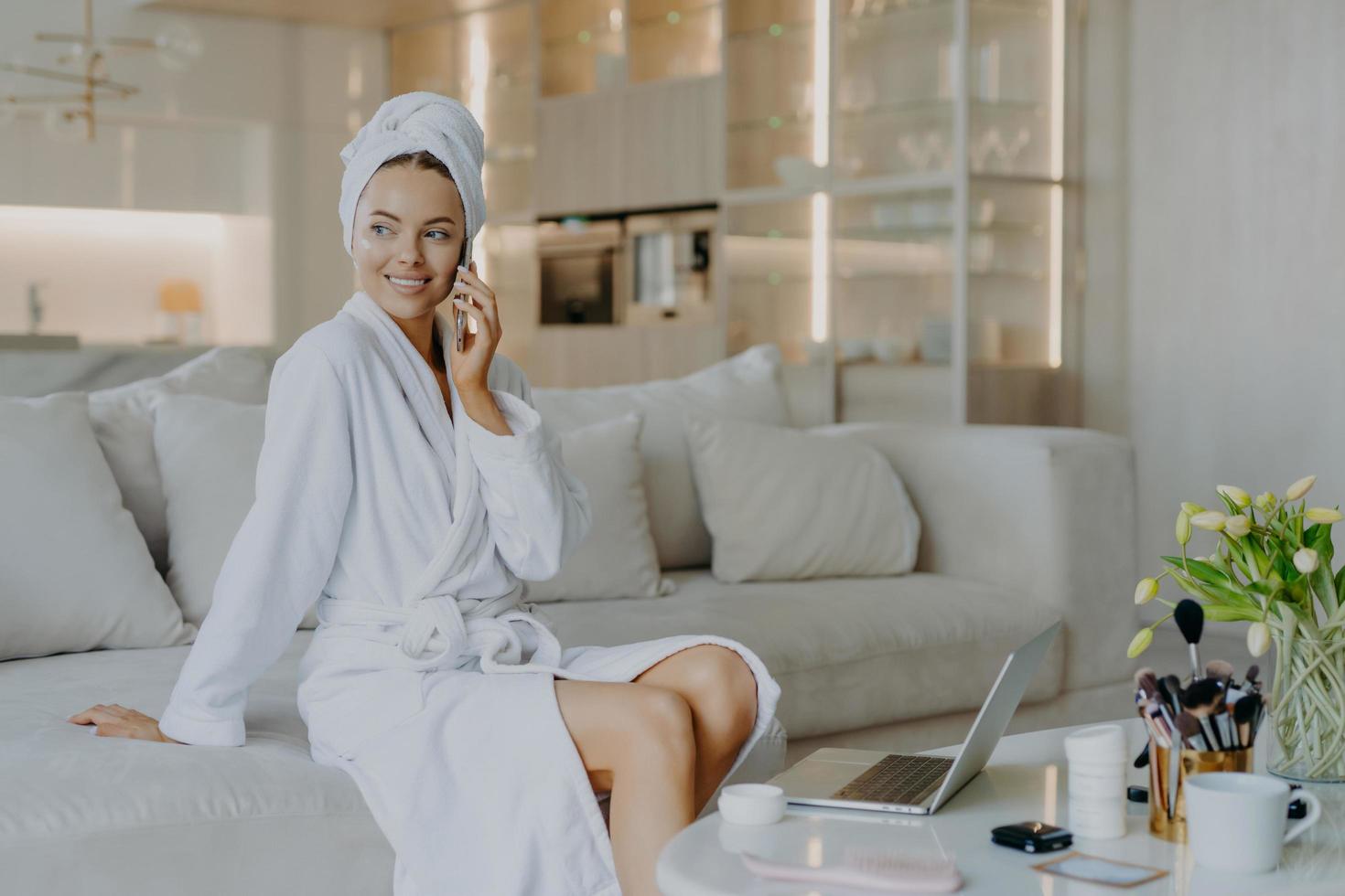 jolie femme réfléchie avec une peau saine un teint bien soigné parle via un téléphone portable bénéficie d'une atmosphère domestique chaleureuse regarde de côté s'assoit sur un canapé confortable dans le salon applique de la crème sur le visage photo