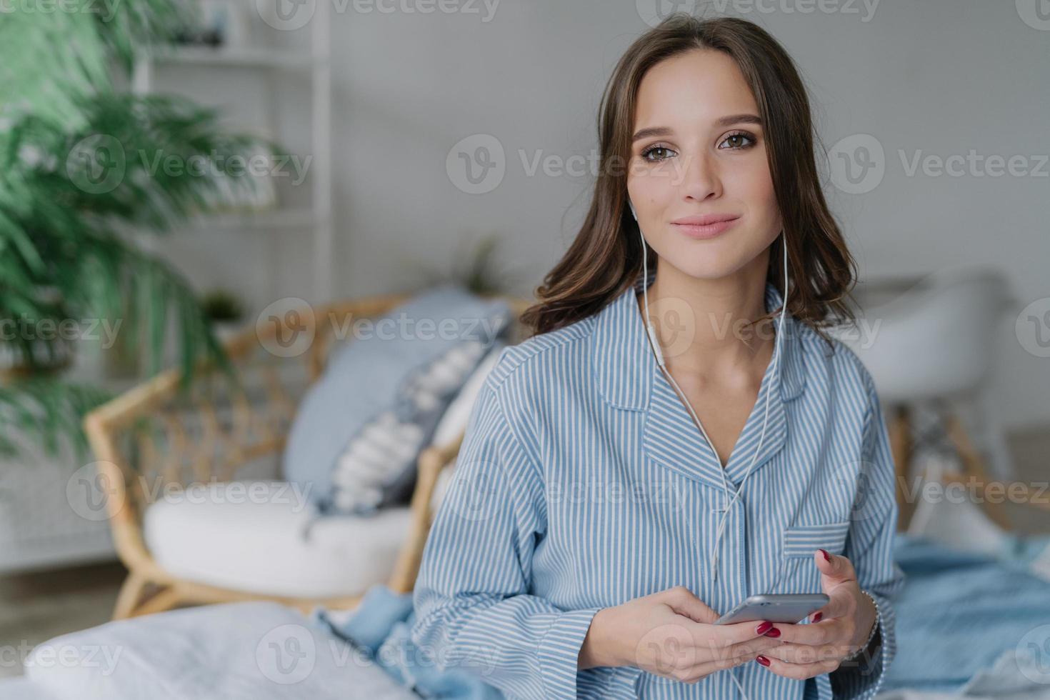 femme séduisante pensive aux cheveux noirs, tient un téléphone portable moderne dans les mains, aime les chansons préférées, utilise un smartphone, pose contre l'intérieur domestique. cours en ligne sur les montres féminines sur un site Web populaire photo