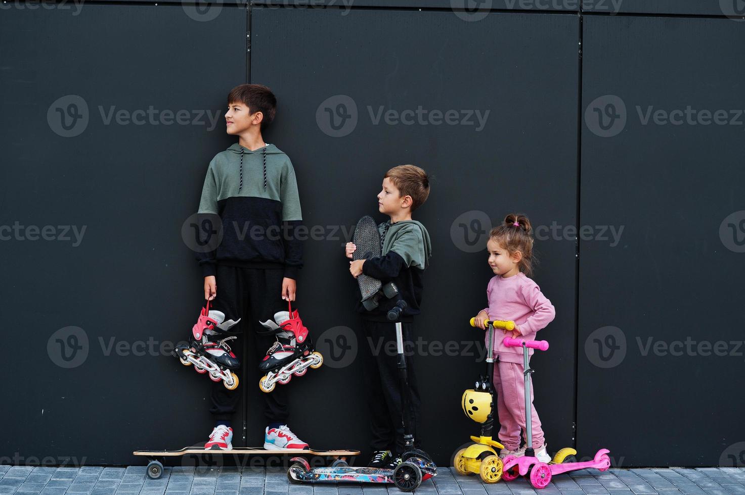 trois enfants en plein air contre un mur moderne noir. la famille sportive passe du temps libre à l'extérieur avec des scooters et des patins. photo