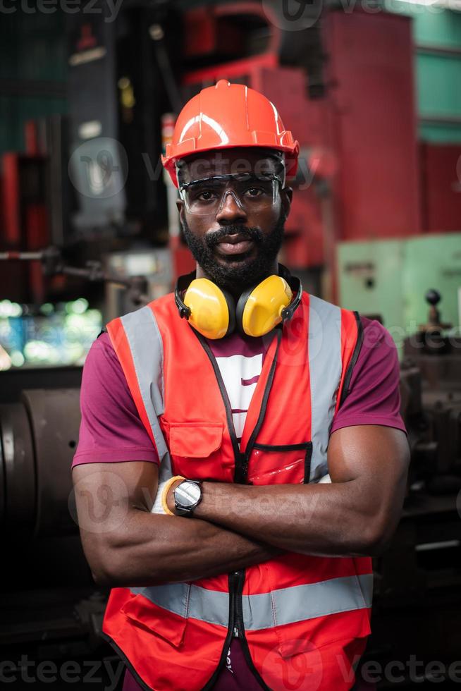 portrait d'un travailleur afro-américain aux yeux sobres et déterminés avec une barbe négligée en uniforme de sécurité portant un casque, des lunettes, un gilet et des gants dans l'usine du fabricant industriel. photo