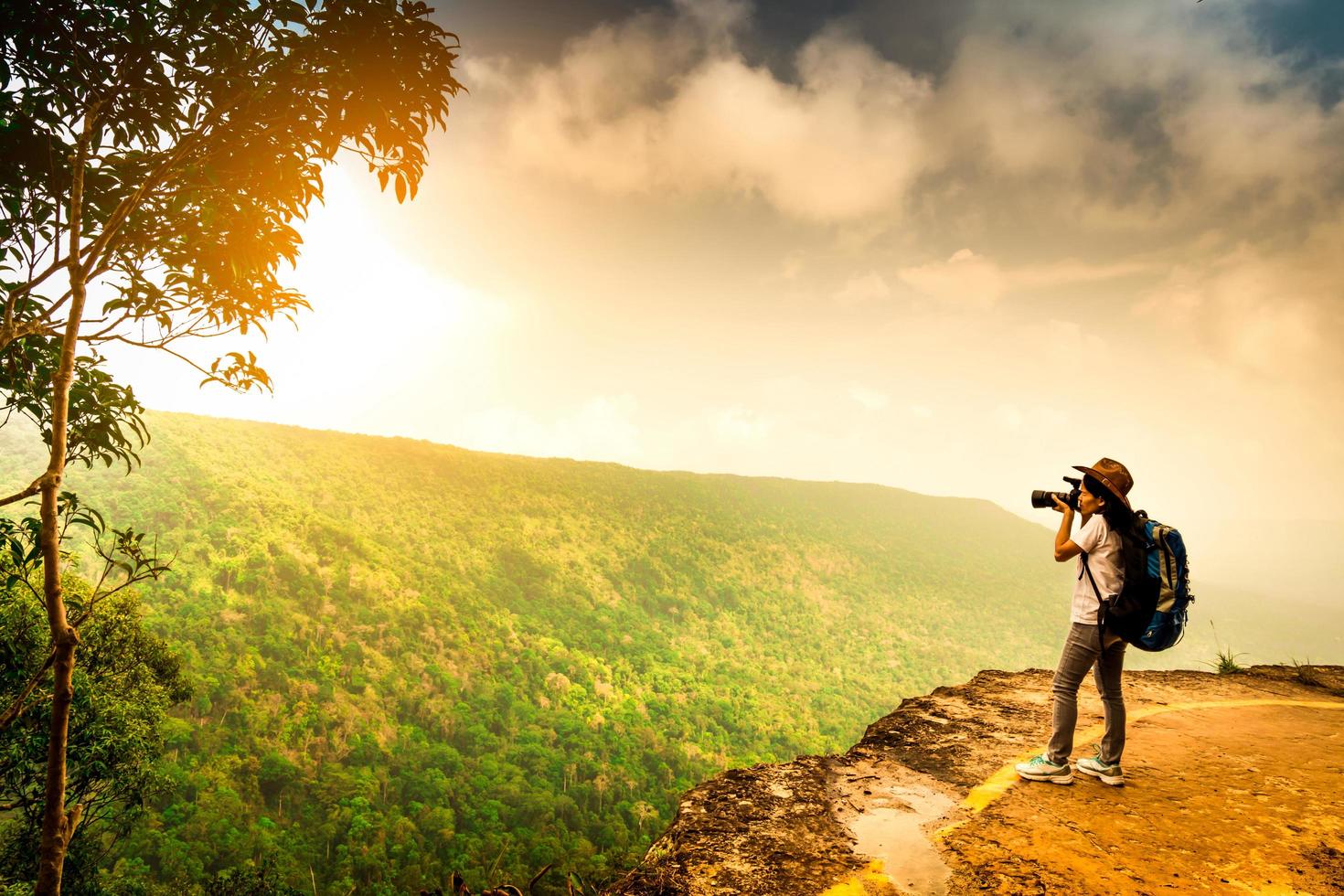 jeune femme voyageant avec sac à dos, chapeau et support de caméra sur la falaise de la montagne. femme photographe prenant des photos de la forêt tropicale, du ciel et des nuages pendant ses vacances. une femme asiatique voyage seule.