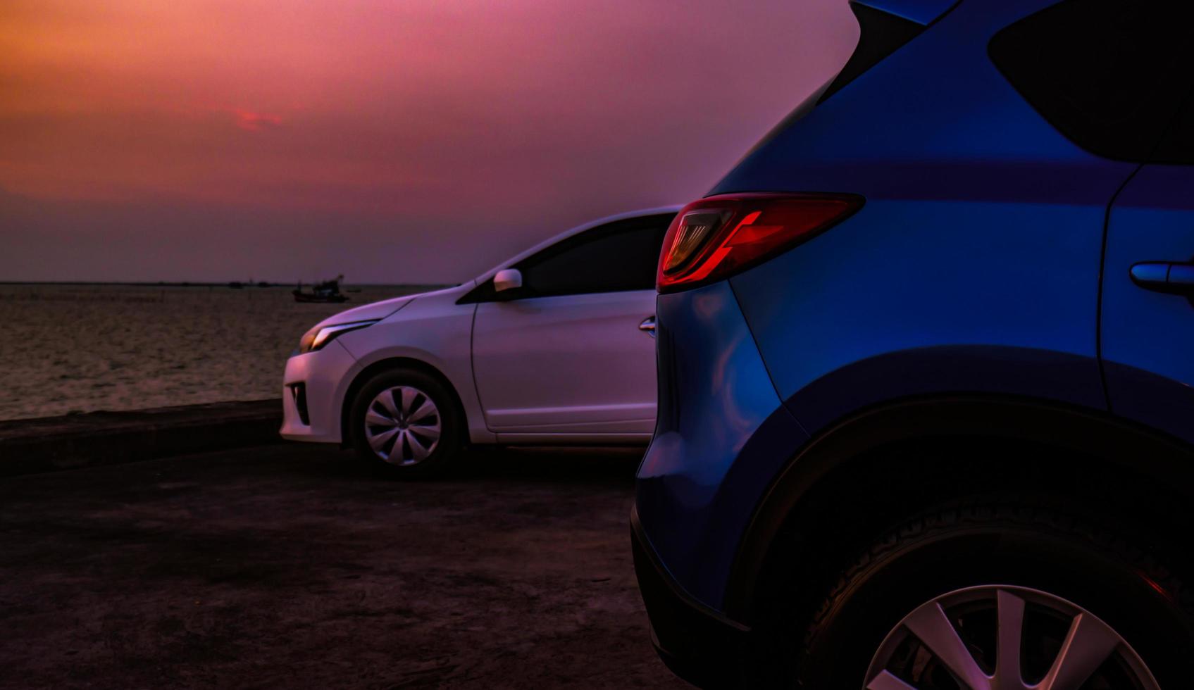 gros plan suv bleu de luxe et voiture blanche garée sur un parking en béton à côté de la plage le soir avec un beau ciel de coucher de soleil violet. voyage sur la route en vacances d'été sur la plage de la mer tropicale. photo