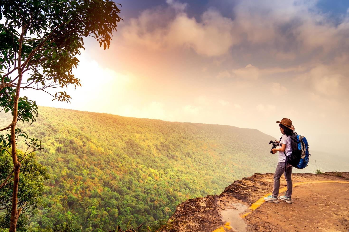 jeune femme voyageant avec un chapeau de sac à dos et un support de caméra au sommet de la falaise de la montagne en regardant une belle vue sur les bois et le ciel après la pluie pendant ses vacances. une femme asiatique voyage seule. photo