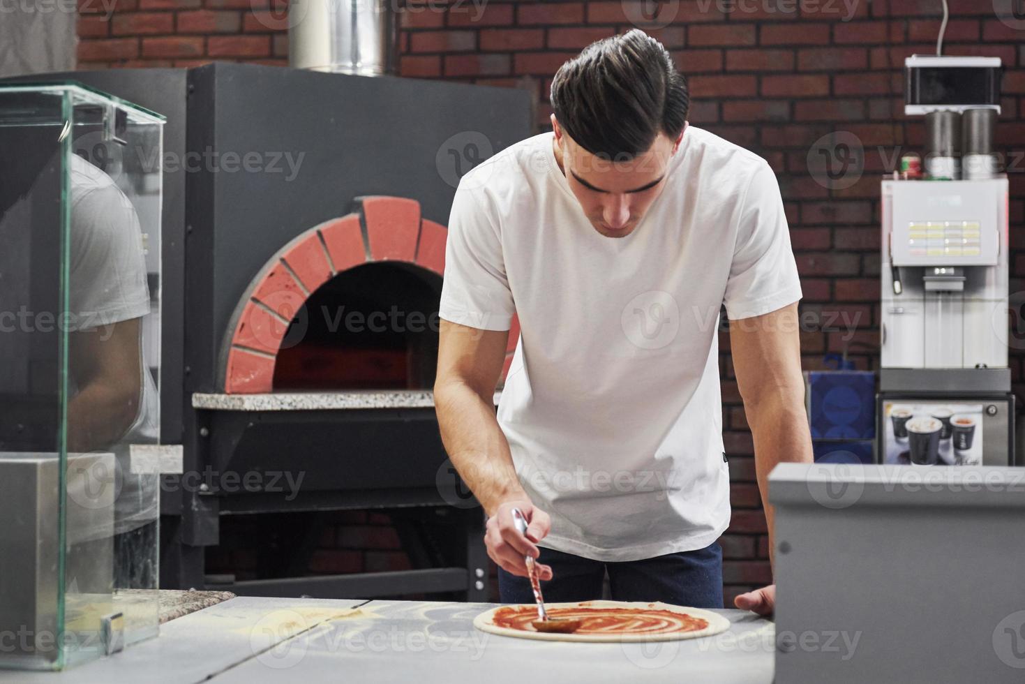 vue de face. boulanger en chemise blanche mettant de la sauce pour faire de délicieuses pizzas pour une commande au restaurant photo