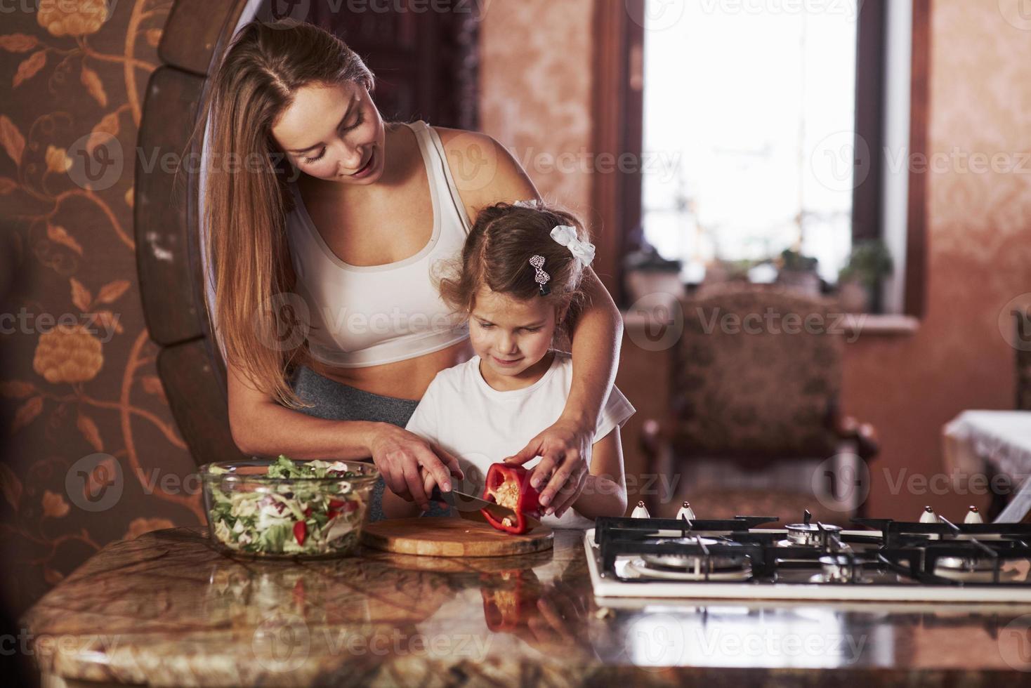 vous pouvez facilement le faire. jolie jeune femme debout dans la cuisine moderne près de la cuisinière à gaz et apprend à sa fille à préparer la nourriture photo