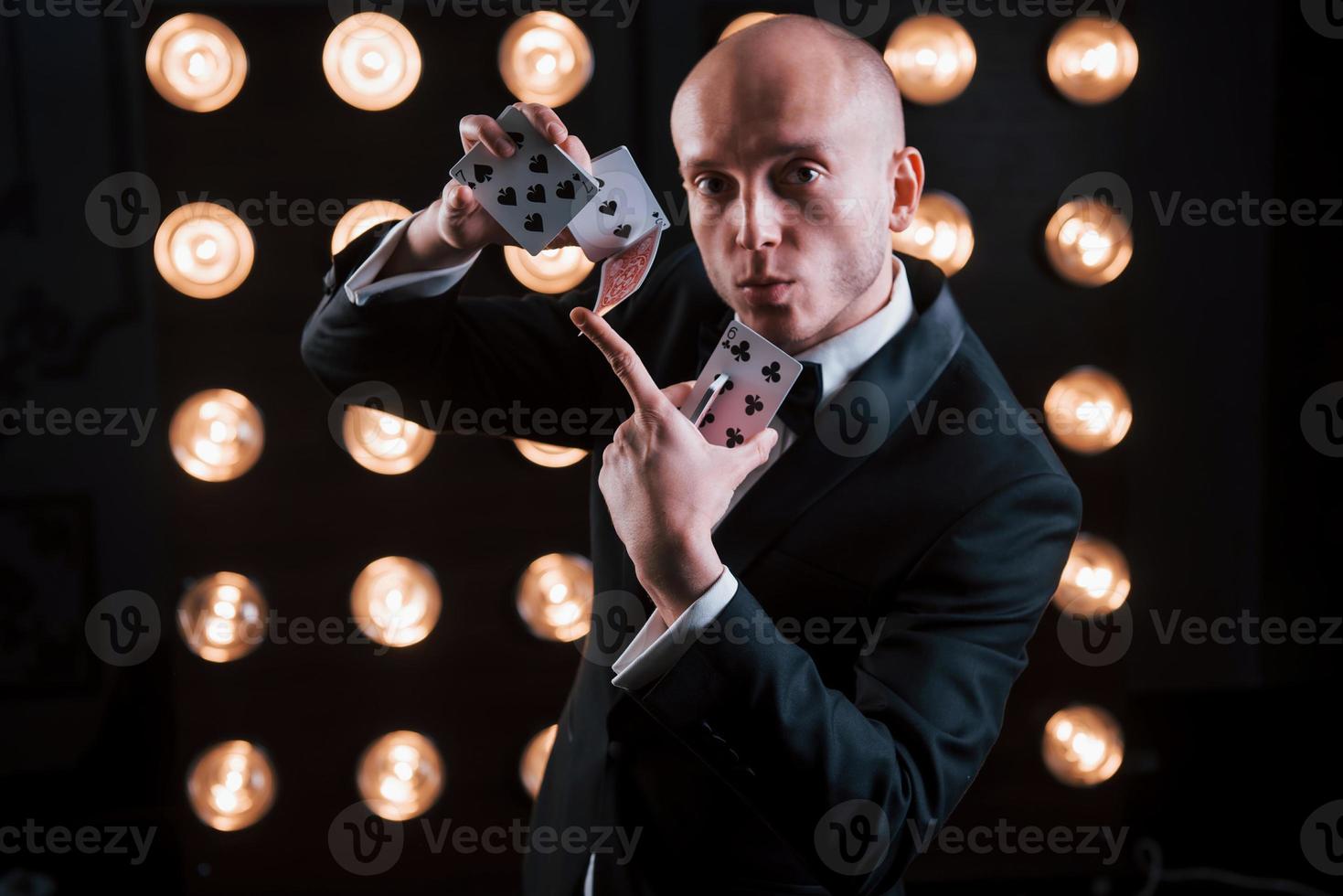 aime son métier d'illusionniste. magicien en costume noir et avec des cartes à jouer debout dans la pièce avec un éclairage spécial dans les coulisses photo