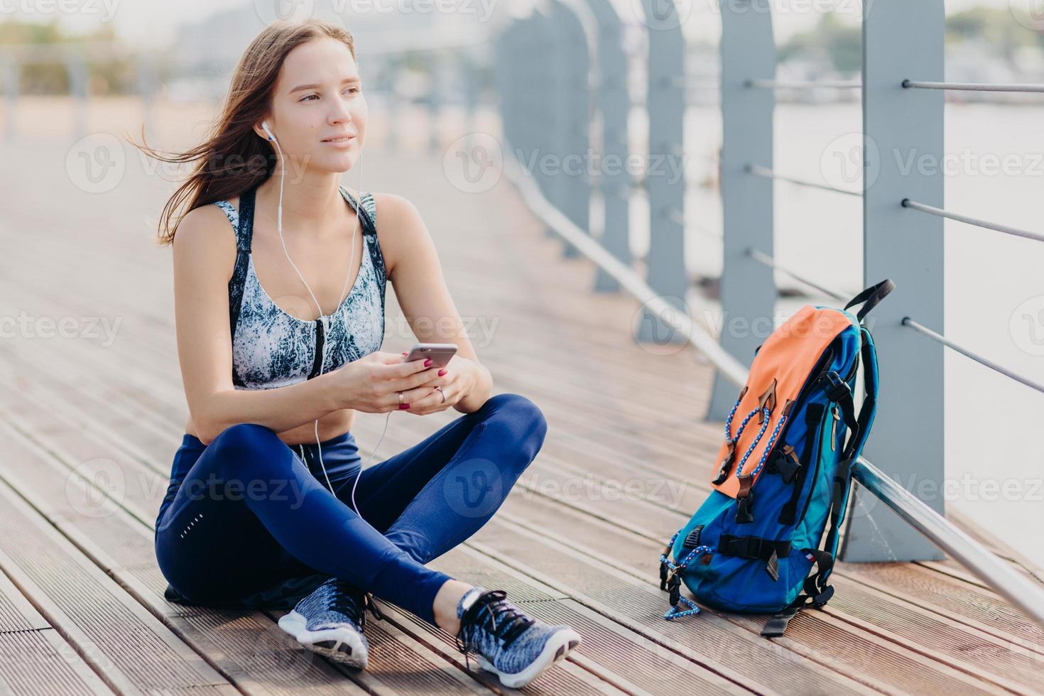 une femme séduisante et rêveuse porte un haut et des leggings, pense à quelque chose tout en écoutant de la musique, tient un téléphone portable moderne, un sac se tient près, respire de l'air frais le matin. concept de loisirs et de style de vie photo