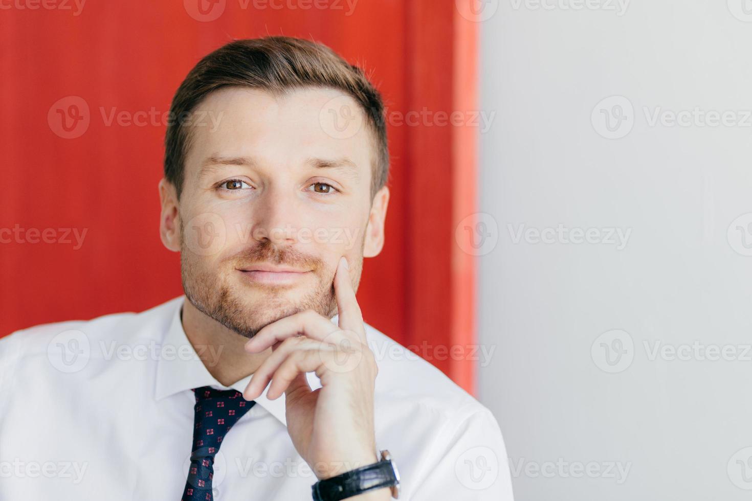 photo de tête d'un jeune employé de bureau mal rasé sérieux tient le menton, regarde directement la caméra, vêtu d'une chemise blanche avec une cravate élégante, pose sur fond rouge. homme d'affaires pose en studio