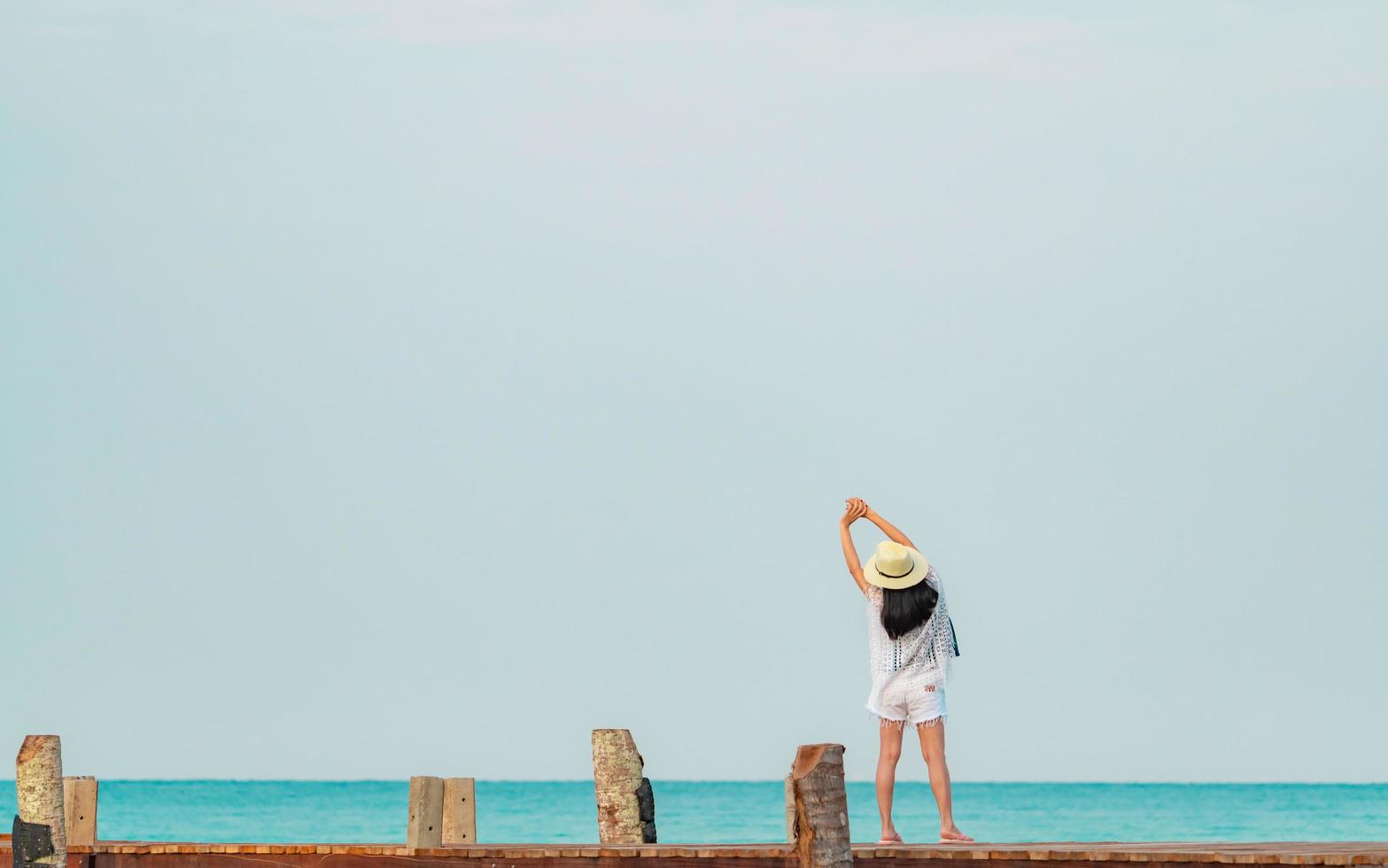 vue arrière d'une jeune femme asiatique heureuse dans un style décontracté et un chapeau de paille se détendre et profiter de vacances à la plage paradisiaque tropicale. fille debout sur la jetée en bois de la station balnéaire à la mode des vacances d'été. photo