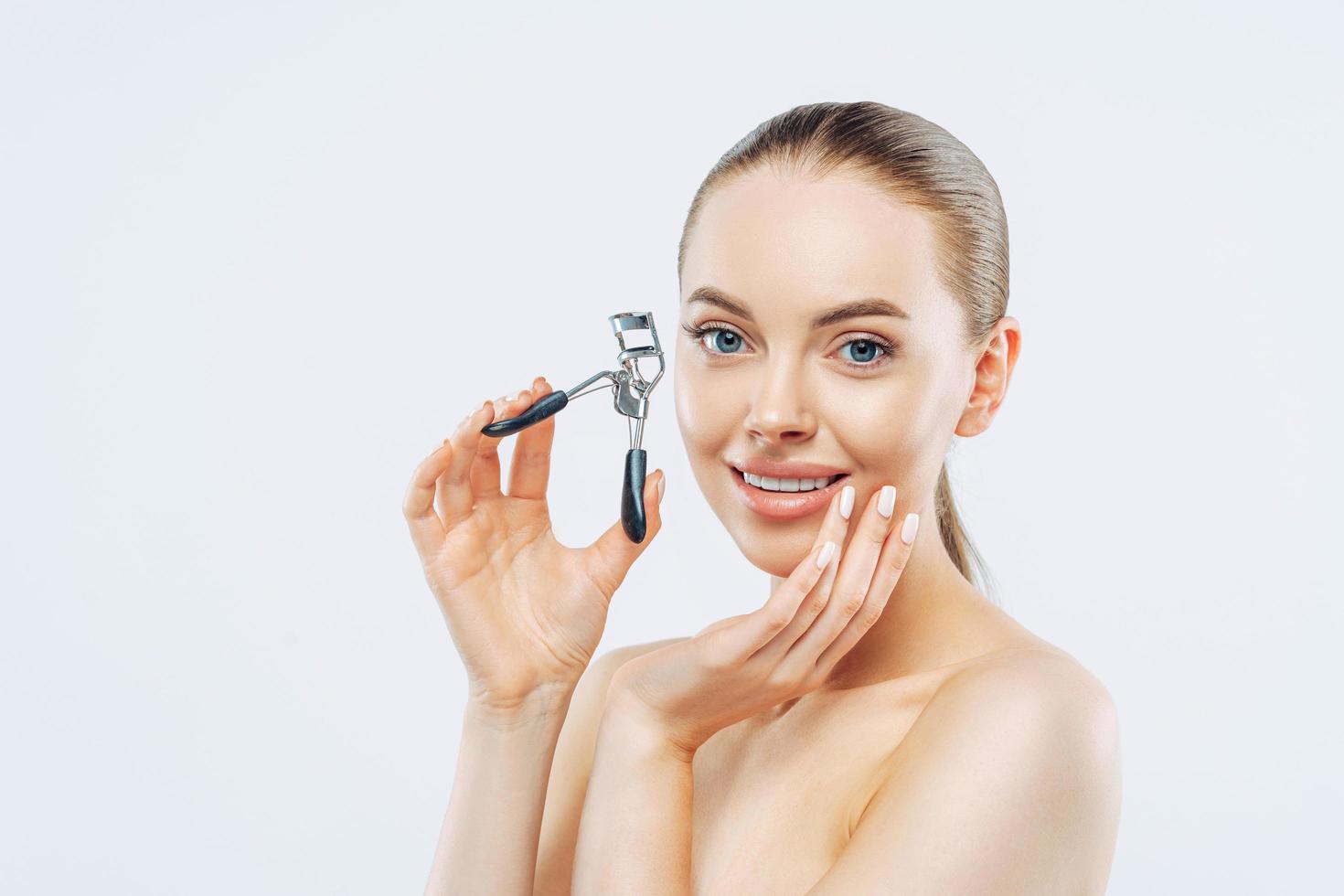 photo en studio d'une jeune femme brune au sourire tendre, utilise un recourbe-cils, a une peau douce et lisse, se tient nue, a un maquillage naturel, touche le visage, pose sur fond blanc. haute résolution