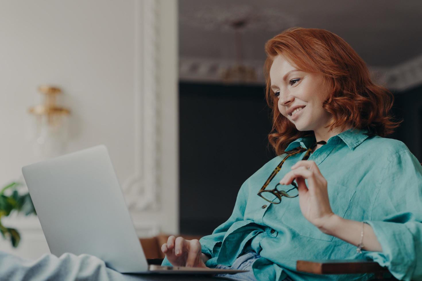 vue latérale d'une jolie femme rousse concentrée sur un travail à distance, prépare une publication pour une page Web, regarde avec plaisir un ordinateur portable, s'assoit dans un appartement moderne, travaille à distance, tient des lunettes photo