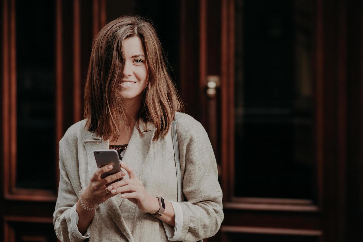 séduisante jeune blogueuse souriante envoie des SMS à ses abonnés, a du temps de récréation, est de bonne humeur, porte un imperméable blanc à la mode, se tient à l'extérieur près des portes. notion de communication photo