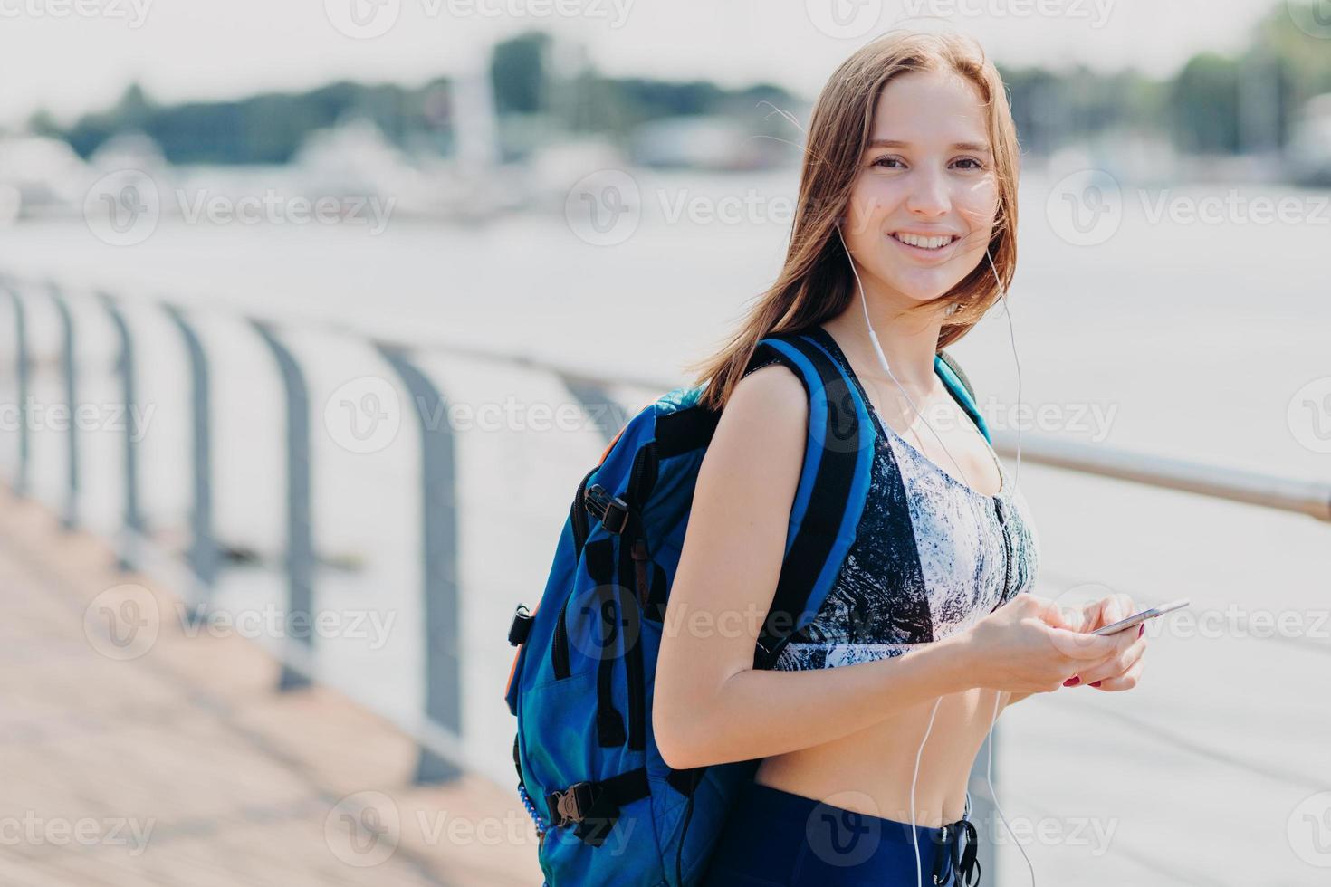 belle femme souriante avec une expression heureuse, porte un haut décontracté, porte un sac, tient un téléphone portable dans les mains, aime la communication en ligne avec des amis, est toujours en contact, écoute de la musique populaire photo