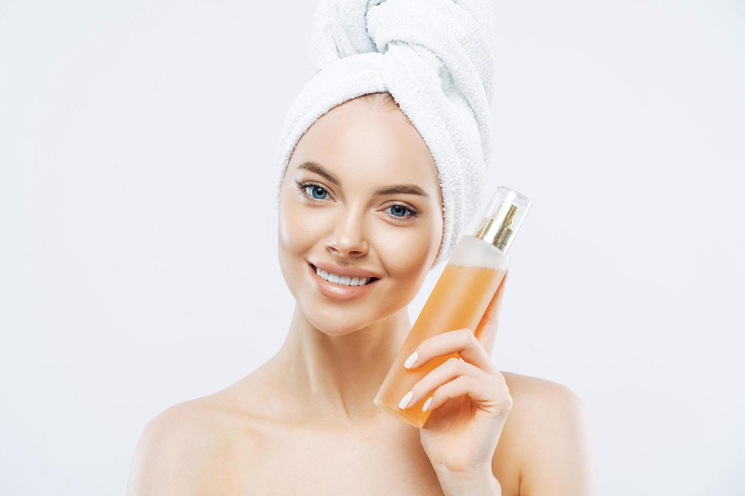 jeune femme au sourire positif, tient une grande bouteille de parfum aromatique à l'odeur de fleur, a un teint soigné, une peau saine, porte une serviette de bain sur la tête, se tient torse nu isolé sur fond blanc photo