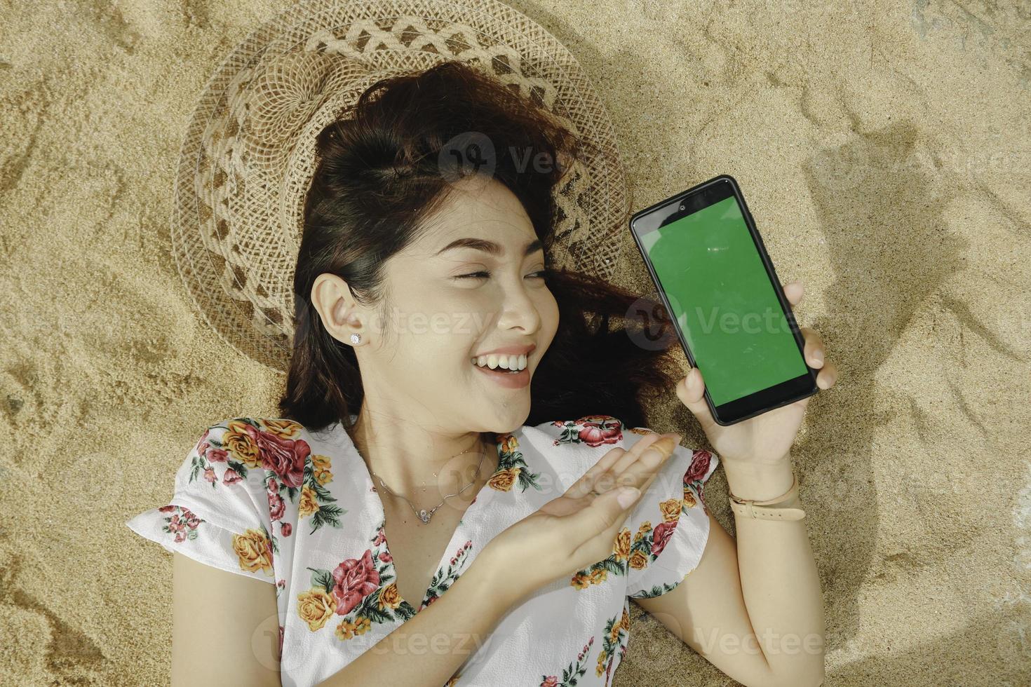 une belle jeune femme asiatique allongée sur le sable tout en montrant l'écran du téléphone photo