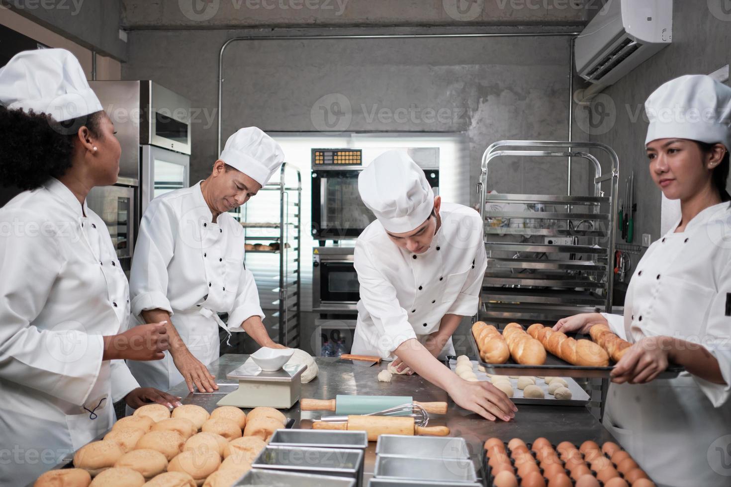 équipe gastronomique professionnelle multiraciale, quatre chefs en uniformes et tabliers de cuisinier blancs pétrissent la pâte à pâtisserie et la farine, préparent le pain et les aliments de boulangerie, cuisent au four dans la cuisine du restaurant en acier inoxydable. photo