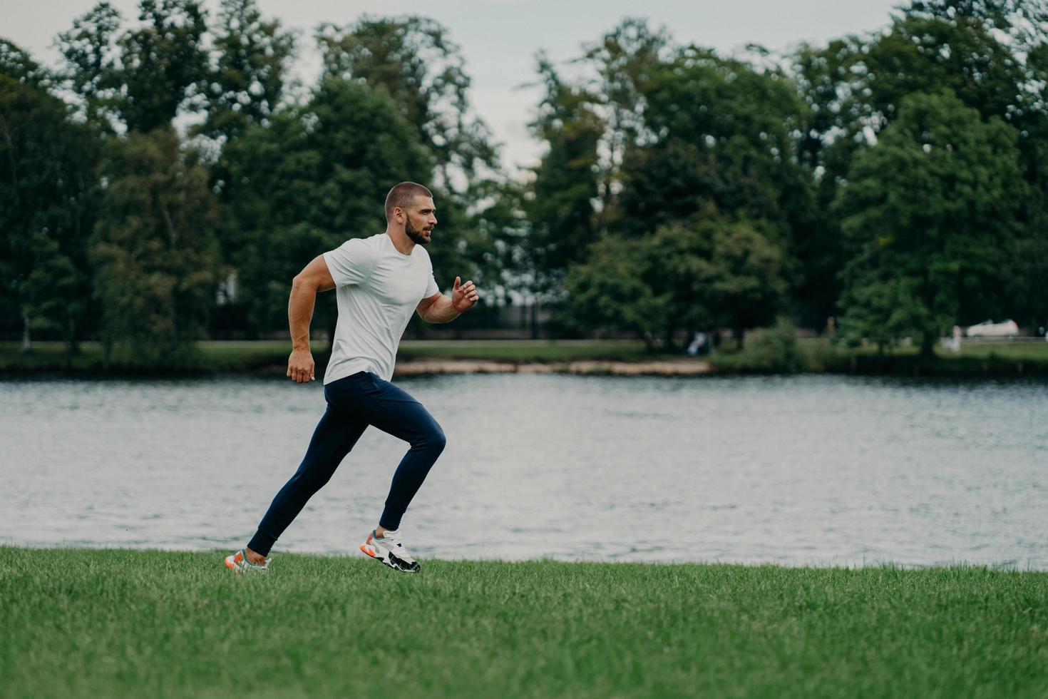 photo horizontale d'un bel homme barbu actif qui court en se faisant photographier en mouvement, court contre la rivière et la belle nature, se prépare pour le marathon. concept de sport, de bien-être et d'entraînement