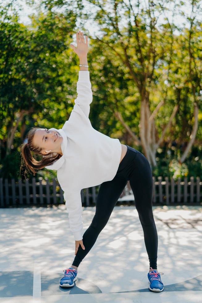 photo verticale d'une sportive heureuse avec une queue de cheval peignée se penche de côté garde un bras levé fait des exercices à l'extérieur porte des leggings à capuche blancs des formateurs se détend sur la nature a un entraînement de fitness actif