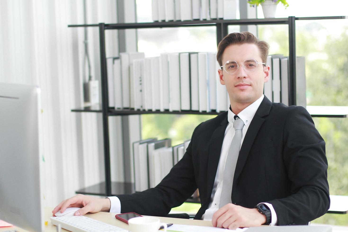 bel homme d'affaires en costume noir porte des lunettes et heureux de travailler avec un ordinateur, un ordinateur portable, un ordinateur portable et un smartphone sur le bureau dans un bureau moderne photo