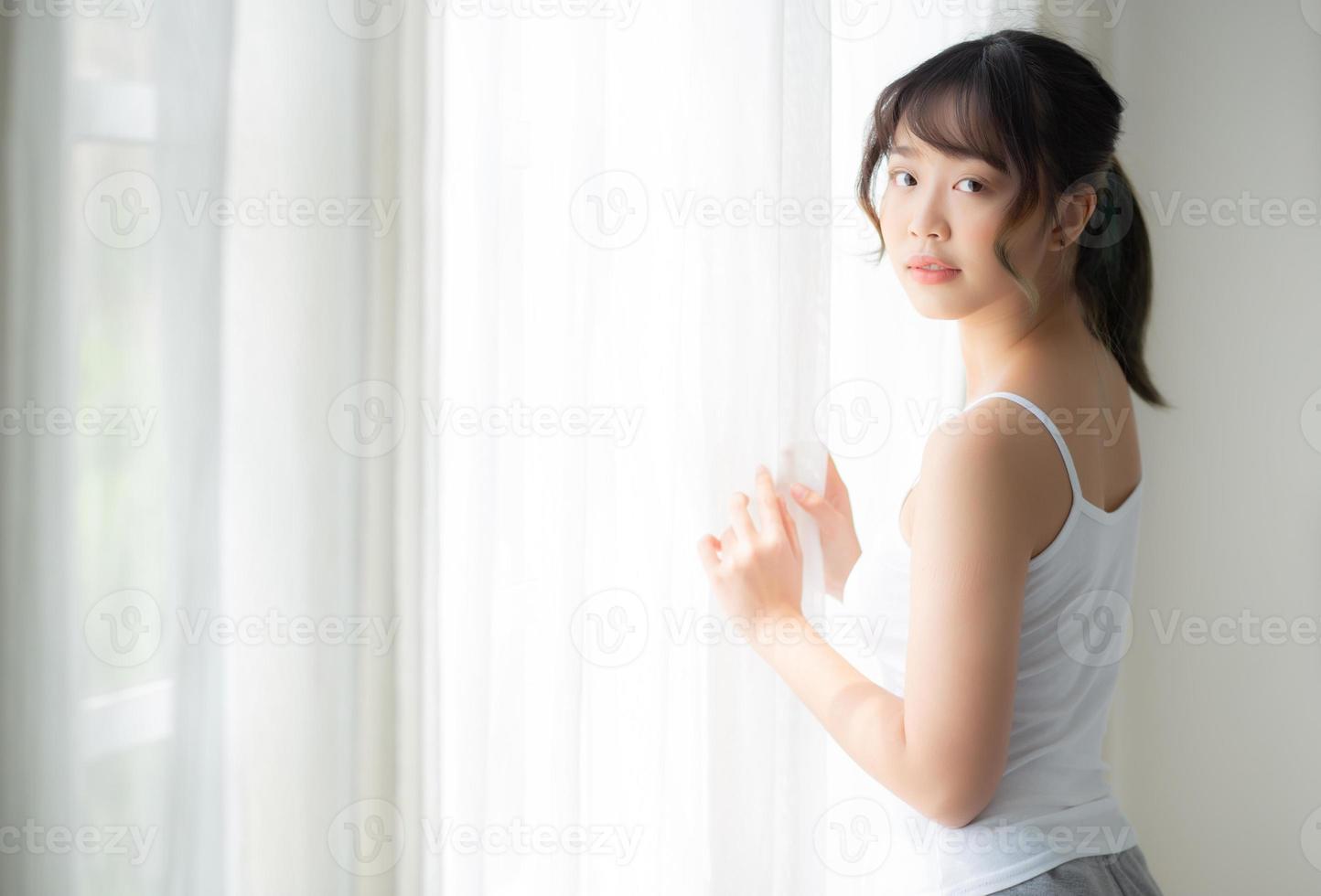 portrait d'une belle jeune femme asiatique debout à la fenêtre et souriant tout en se réveillant avec la lumière du soleil le matin, fille heureuse avec frais et gai, style de vie et détente pour le concept de bien-être. photo