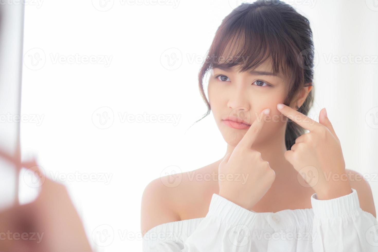 belle jeune femme asiatique regardant le miroir est une acné, un traitement de zit, un problème de fille beau visage, une beauté parfaite avec le bien-être dans la chambre à la maison avec un concept de soins de la peau et de soins de santé. photo