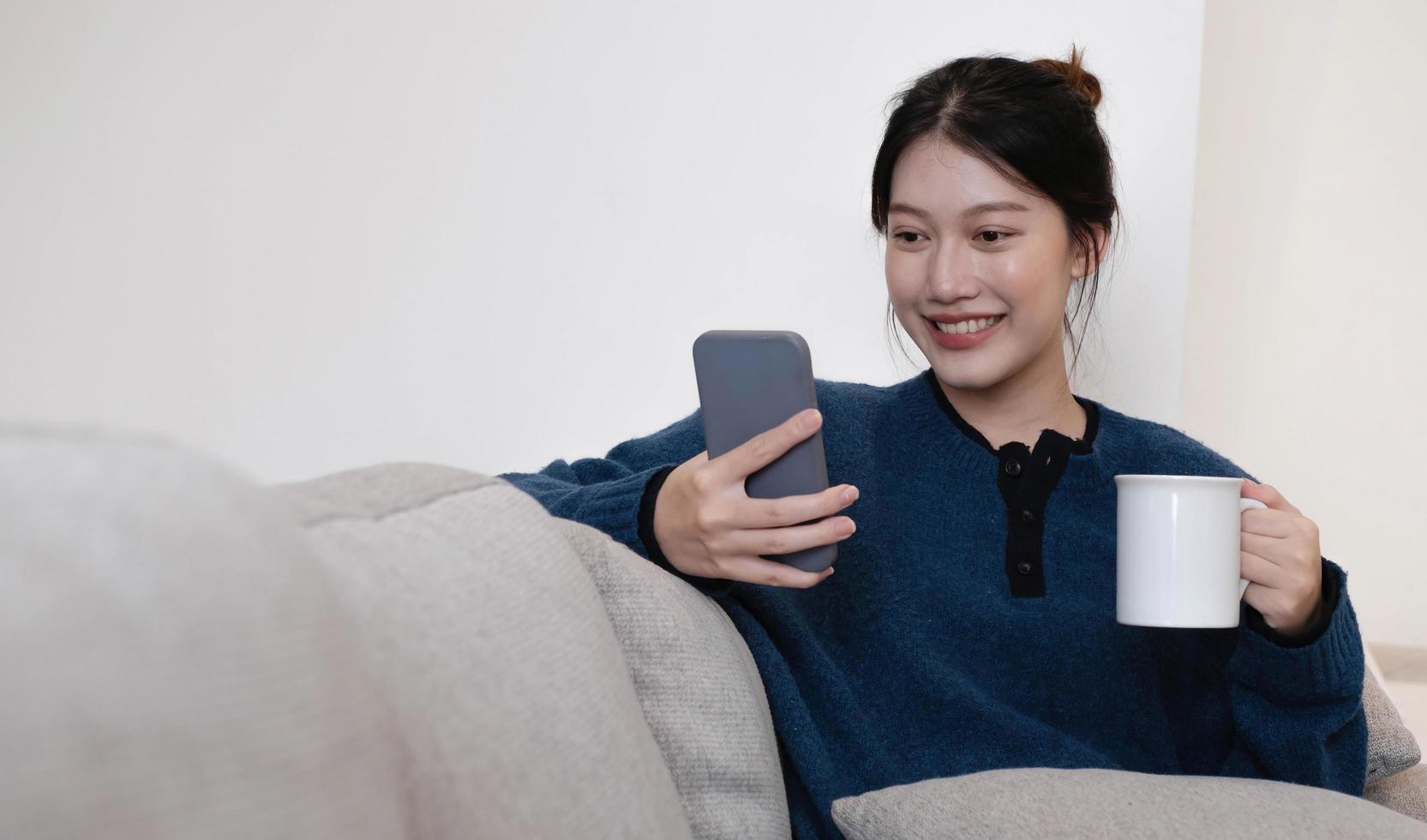portrait d'une femme asiatique souriante assise sur un canapé et buvant du café en regardant un smartphone. photo