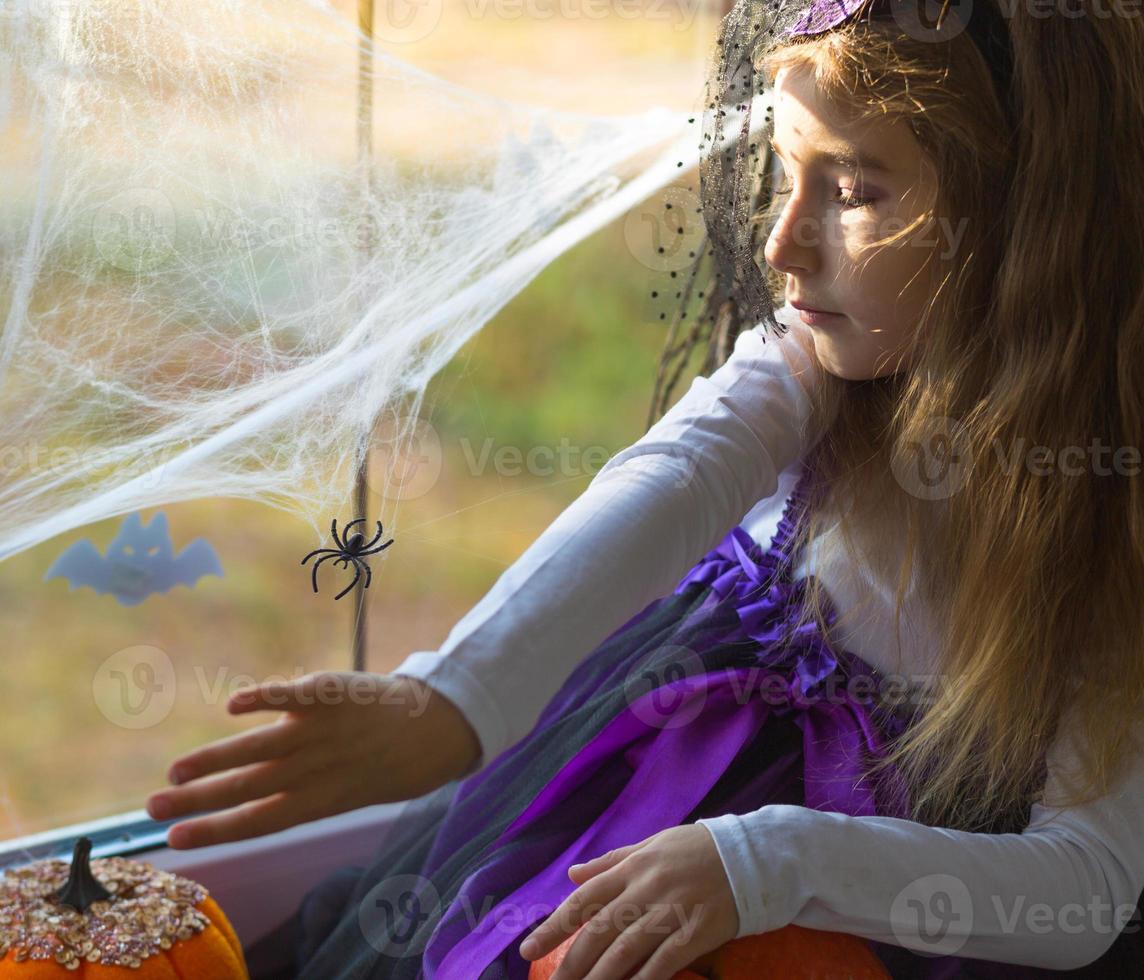 une fille en costume de sorcière sur le rebord de la fenêtre près de la fenêtre joue avec une toile d'araignée et une araignée, décorant la maison pour les vacances d'halloween. paysages terribles, peur et horreur, citrouilles photo