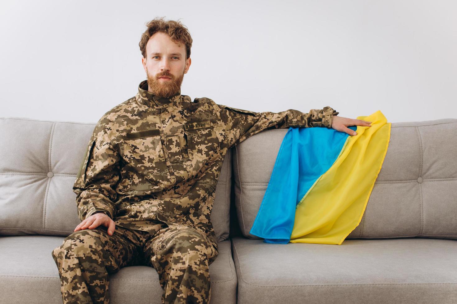 portrait d'un jeune soldat patriote ukrainien émotif en uniforme militaire assis sur le bureau sur un canapé avec un drapeau jaune et bleu. photo