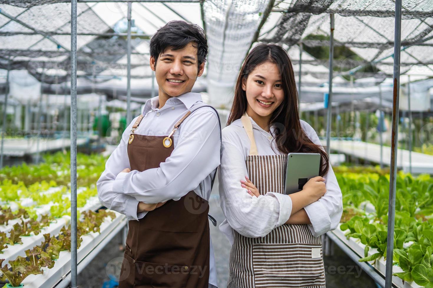 nouvelle génération de deux jeunes agriculteurs asiatiques souriants travaillant dans une ferme hydroponique de légumes, propriétaires de potagers hydroponiques prospères debout dans une plantation de serre pour une alimentation saine photo