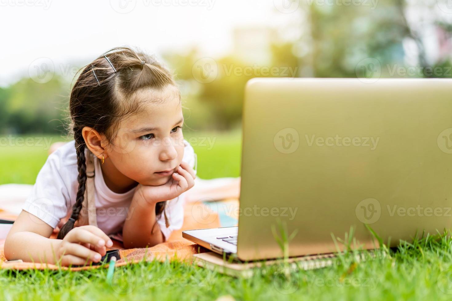 heureuse petite fille souriante, parlant, apprenant et s'amusant en regardant un ordinateur portable dans le parc d'été. apprentissage du concept d'éducation en ligne photo