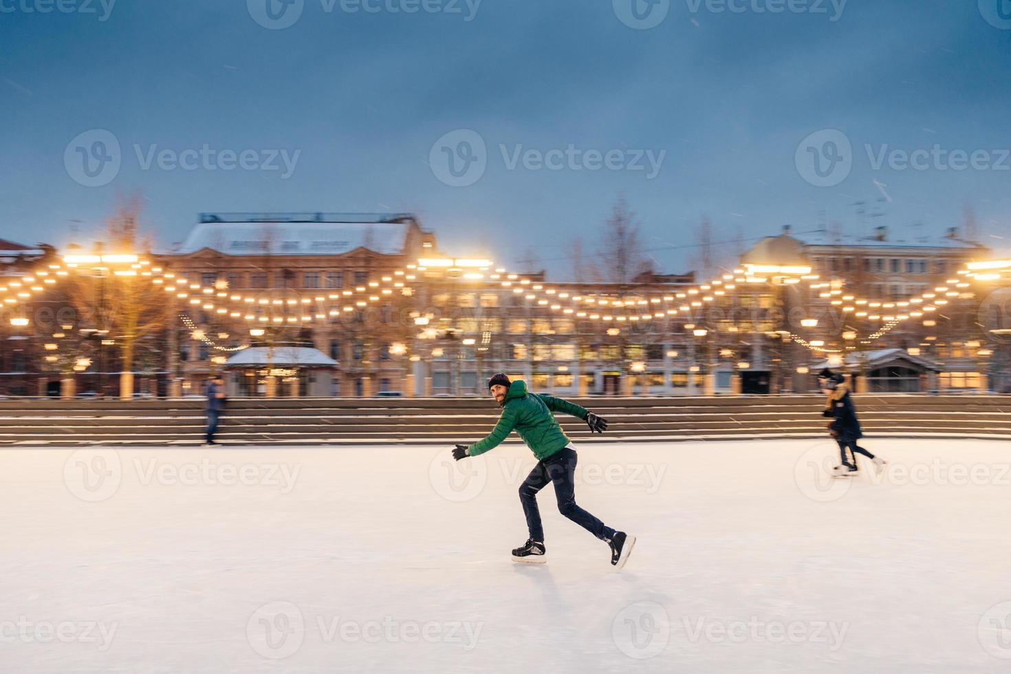 un homme actif s'amuse dans un parc extérieur sur une patinoire décorée de guirlandes, montre ses talents de patineur, fait des mouvements rapides sur des patins, est confiant, aime les activités de plein air hivernales photo