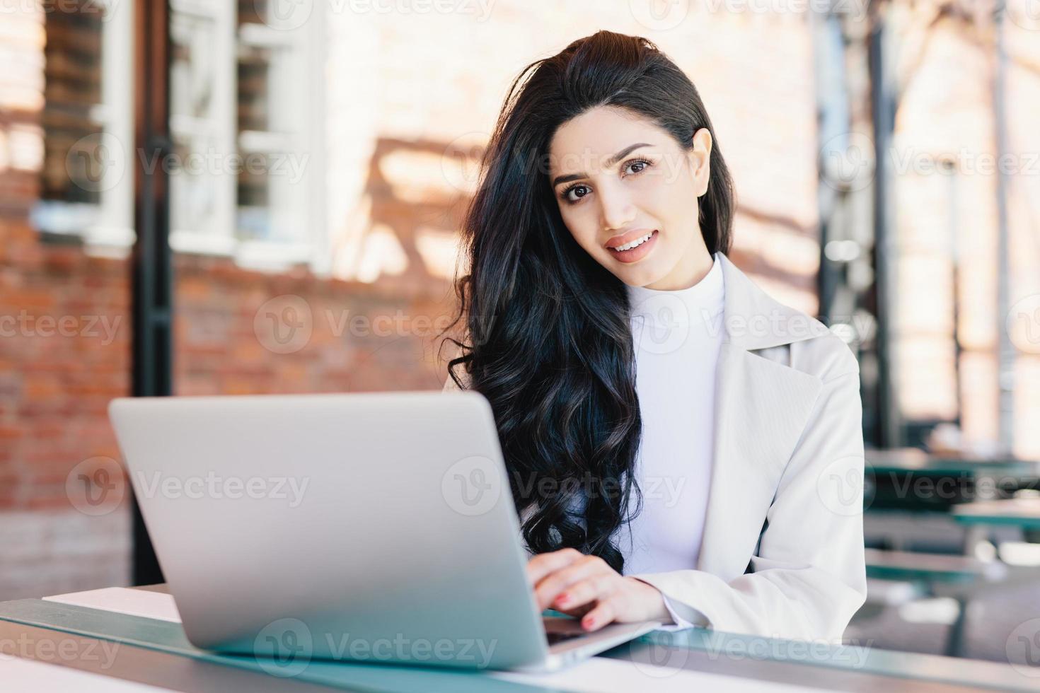 concept de technologie et de communication. femme d'affaires européenne prospère avec une belle apparence travaillant dans un café sur un ordinateur portable ayant une expression souriante regardant à huis clos tout en étant assis à l'extérieur photo