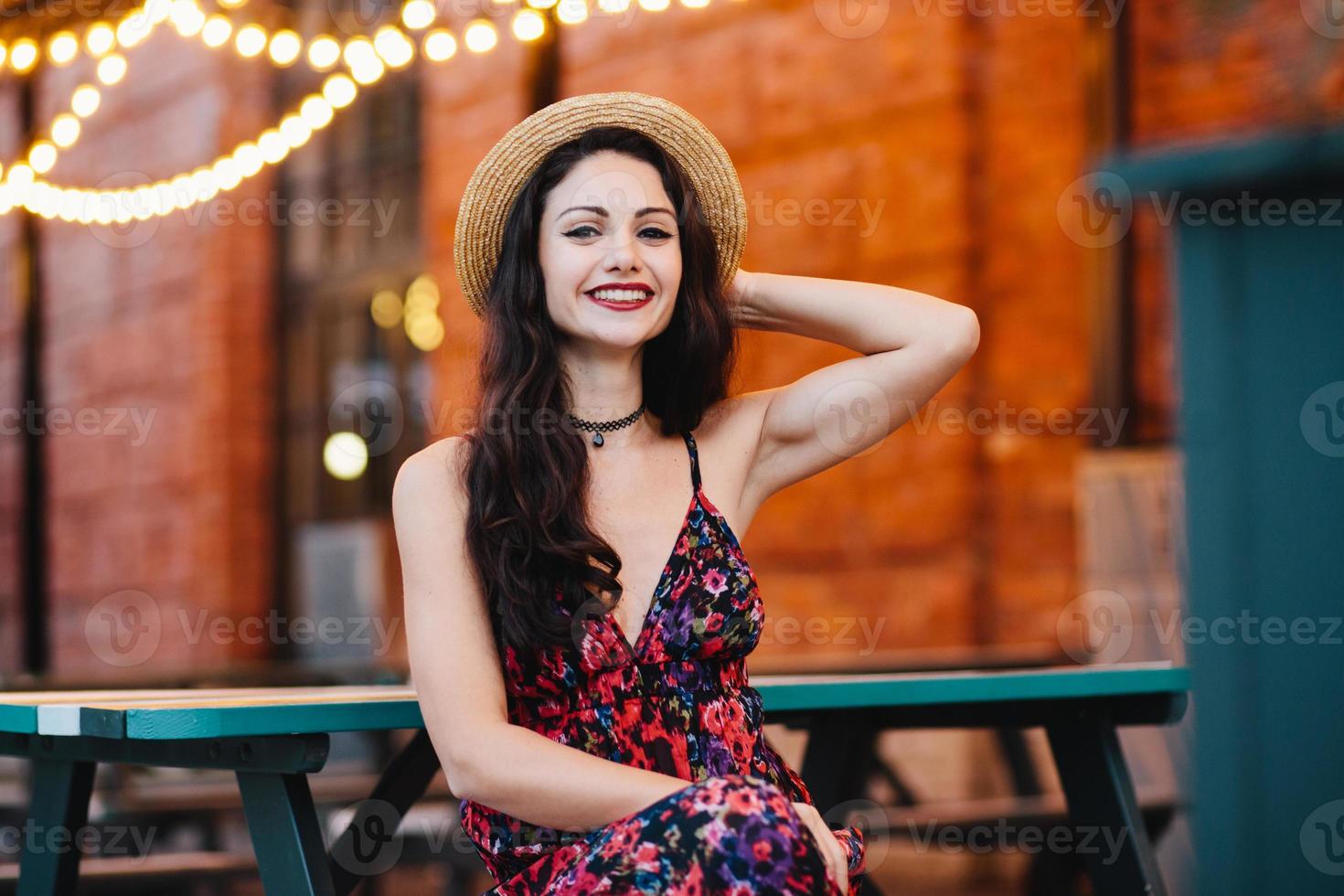 les gens, le bonheur et le concept de mode. jolie femme aux sourcils bien formés, aux yeux noirs et aux lèvres rouges, portant un chapeau et une robe de paille, assise à la cafétéria, admirant la bonne ambiance photo