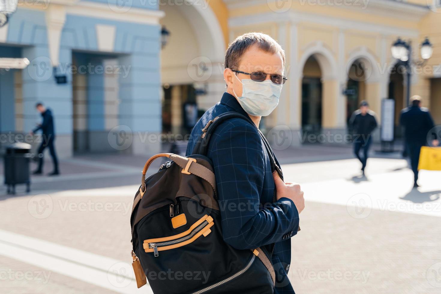 vue arrière de l'homme voyageur porte un masque médical pour la prévention des virus porte un sac à dos sur l'épaule pose dans un lieu public, une gare routière ou ferroviaire, se tient à l'extérieur, protège contre le coronavirus, maladie contagieuse photo