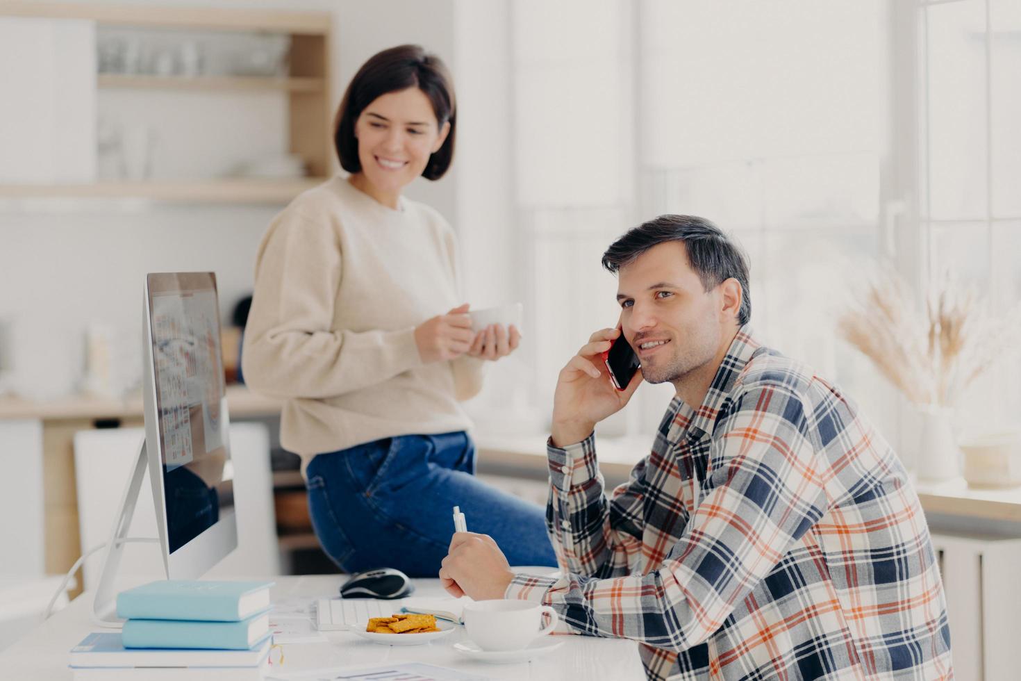 un mari et une femme heureux gèrent les finances à la maison, examinent les comptes bancaires, un homme heureux tient un téléphone portable près de l'oreille, porte une chemise tressée, une femme heureuse est assise sur une table, boit du café aromatique dans une tasse photo