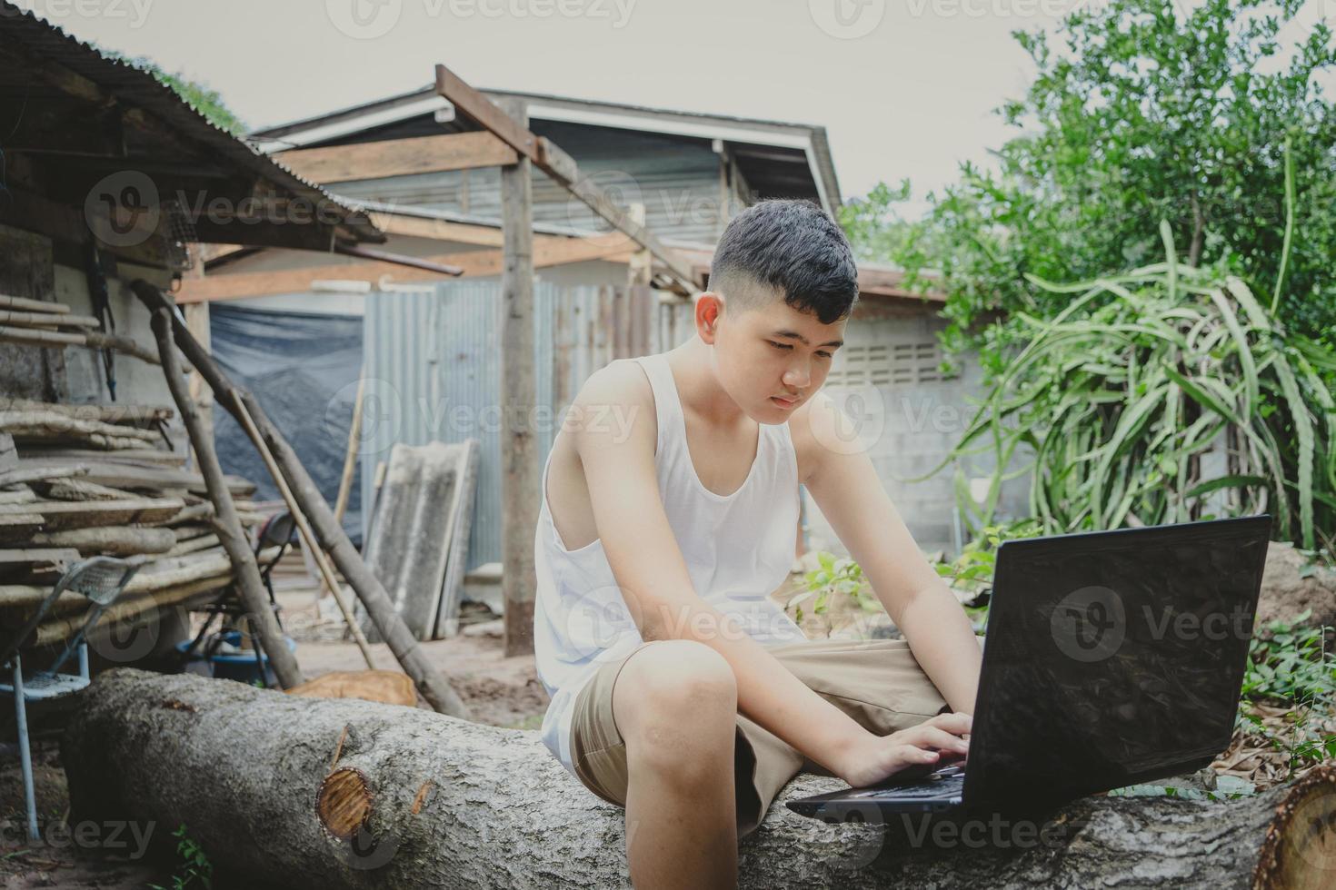 pauvre écolier étudiant en ligne avec un ordinateur portable sur une bûche derrière une vieille maison de campagne. concept d'éducation en ligne dans la campagne et travail à domicile photo