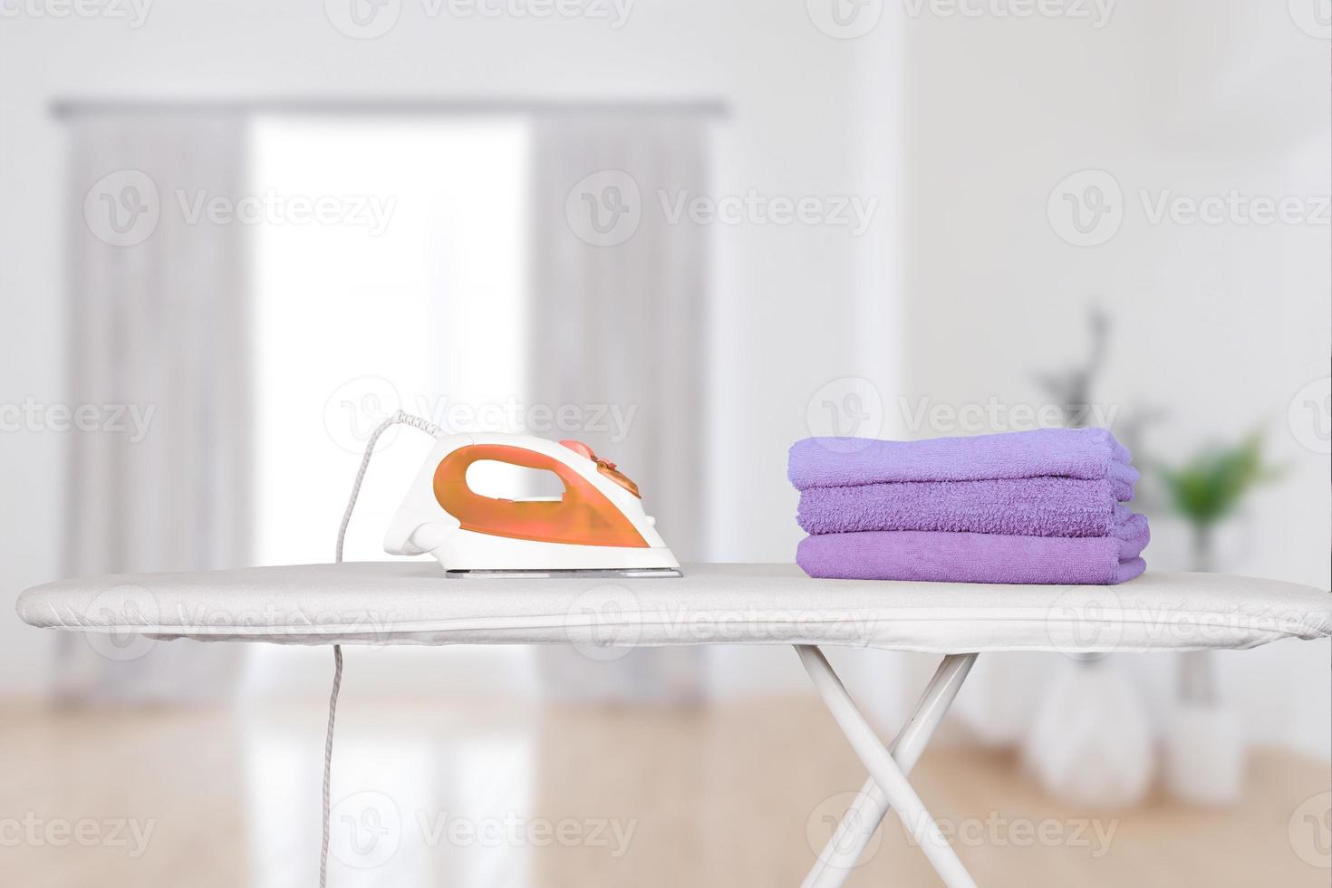 un fer à repasser électrique se dresse sur une planche à repasser et des serviettes de bain pliées en violet très péri, sur fond d'intérieur de maison. arrière-plan flou, mise au point douce. photo