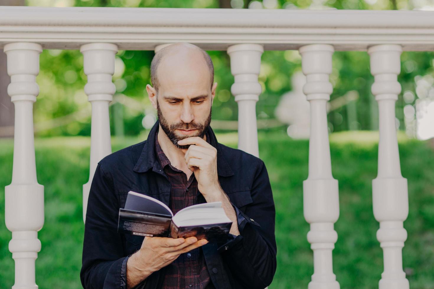 un homme chauve barbu sérieux et concentré tient le menton, lit attentivement le livre, pose à l'extérieur contre un balcon blanc, concentré sur l'intrigue, profite de son temps libre et d'une journée d'été ensoleillée à l'extérieur. repos dans le parc. photo