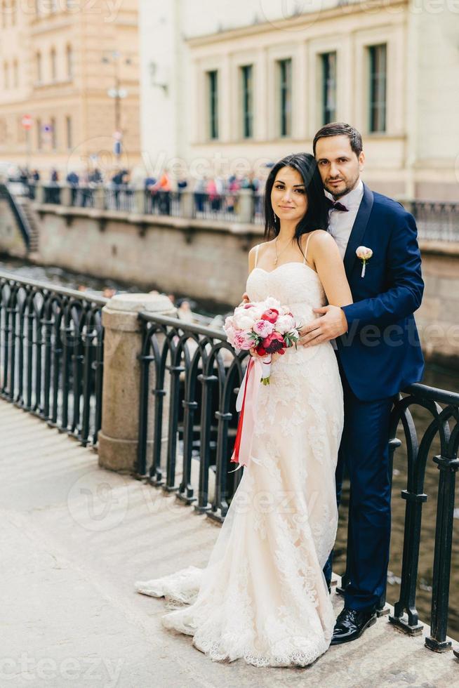 beau marié barbu embrasse sa mariée en robe blanche avec bouquet se tenir ensemble au pont, avoir de la bonne humeur après l'enregistrement de leur mariage. notion de bonnes relations photo