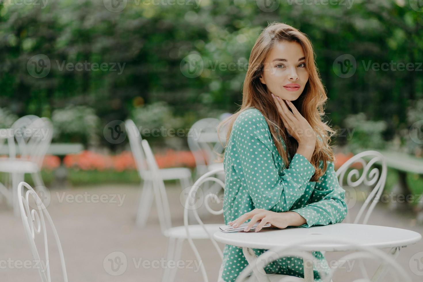 photo latérale d'une belle femme caucasienne en vêtements élégants, porte du maquillage, tient un téléphone portable, passe du temps libre dans un café en plein air, attend un ami. concept de personnes, de style de vie, de repos et de technologie