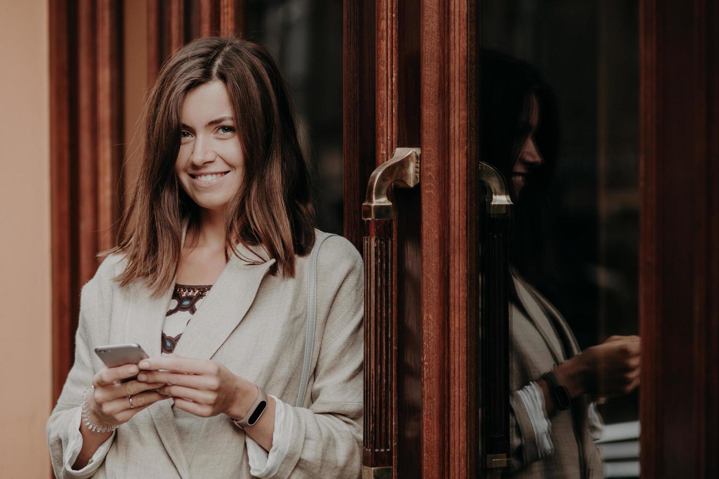 joyeuse femme heureuse aux cheveux raides foncés, utilise un téléphone intelligent, vêtue de vêtements élégants, regarde positivement la caméra, aime la communication en ligne, vérifie l'itinéraire, envoie des messages, lit la notification photo